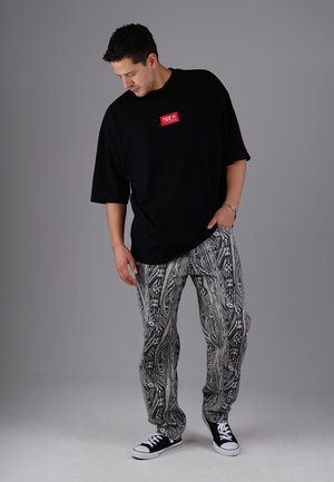 Man wearing a black oversized t-shirt with red logo, black and white patterned pants, black sneakers, looking down with one hand in pocket.