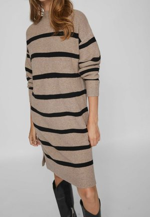 Kobieta w beżowej dzianinowej sukience w czarne poziome paski i czarnych kozakach za kolano stojąca na jednolitym tle.