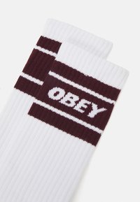 Calcetines blancos con rayas burdeos y el logo "OBEY". Presentan una textura suave y son de longitud hasta la mitad de la pantorrilla, diseñados para ofrecer comodidad y estilo.