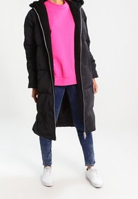 Abrigo de plumas largo y negro con cremallera en la parte delantera, capucha forrada y textura interior suave, llevado sobre un sudadera de color rosa brillante y jeans azules. Zapatillas blancas.