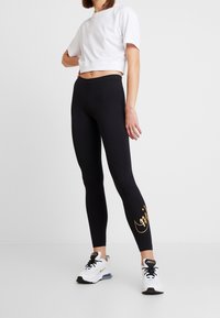 Svarta leggings med en figursydd design, med ett guldaccent på underbenet, i kombination med en vit kortärmad t-shirt och vita sneakers.