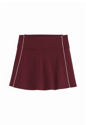 Femme portant un haut blanc sans manches et une jupe bordeaux tenant une raquette de tennis et une veste assortie sur un fond clair uni.