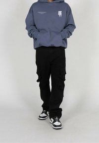 Henkilö, jolla on päällä sininen huppari, jossa on valkoista tekstiä, mustat cargo-housut, joissa on sivutaskut, ja musta-valkoiset lenkkarit, seisoo yksinkertaista taustaa vasten.