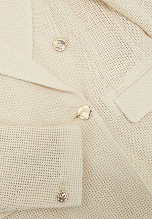 Krémová texturovaná tkanina s třemi lesklými nepravidelnými zlatými knoflíky uspořádanými vertikálně na manžetě a klopě oděvu.