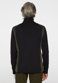 En person med kort grått hår bär en svart och olivgrön långärmad jacka och olivgröna byxor, sedd bakifrån.