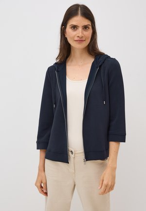Navy zip-up hoodie with a drawstring hood, three-quarter sleeves, and smooth fabric, paired with a light tank top and beige pants.