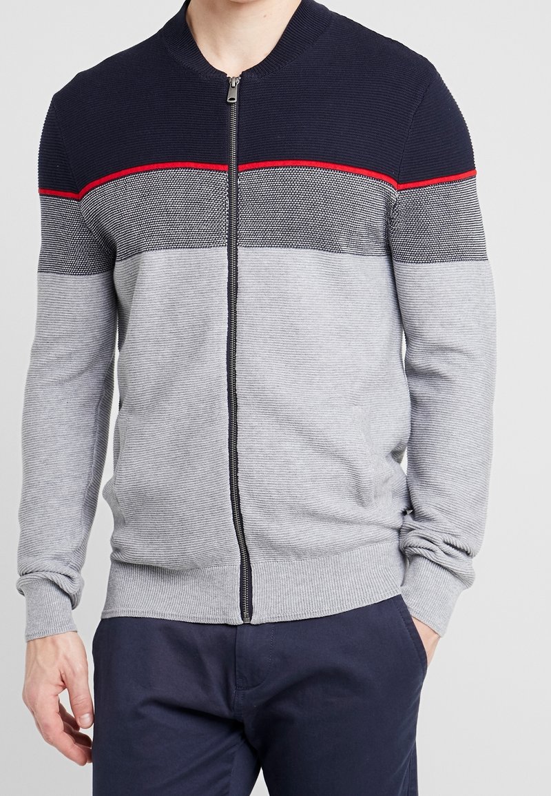 Stickad tröja med marinblå krage, dragkedja framtill och horisontella ränder i grått och svart med en röd accent. Ribbad mudd och nederkant.