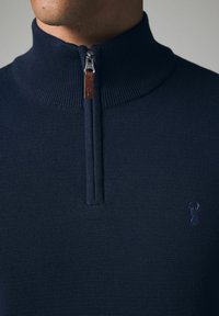 Pulover navy blue cu fermoar, dotat cu un guler înalt, textură nervată și o bandă din piele pentru tragere. Prezintă un logo subtil pe piept.