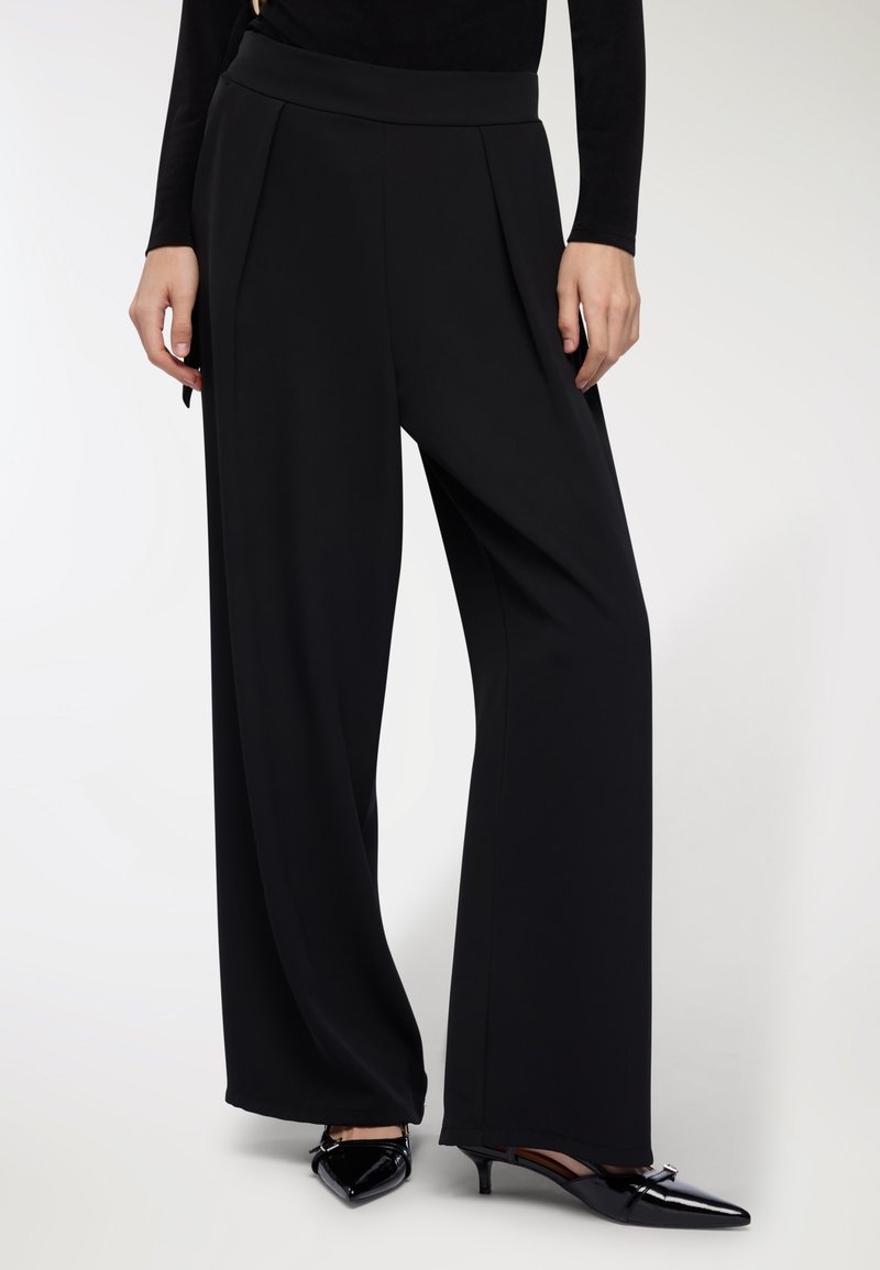 Pantaloni neri a gamba larga con una texture liscia, caratterizzati da una vita alta e pieghe frontali. Abbinati a dei décolleté neri lucidi a punta.