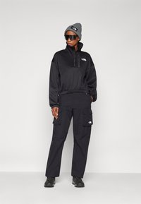 Pullover nero con mezza zip, polsini elasticizzati, pantaloni cargo neri abbinati con tasche, berretto di maglia grigio, occhiali da sole oversize scuri e stivali neri.