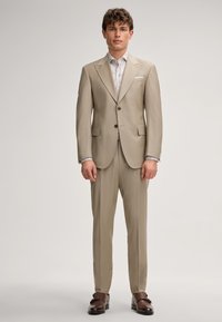 Beige op maat gemaakt kostuum met notch revers, twee knopen en zijzakken. Gepaard met een met patroon shirt en bruine dresschoenen.