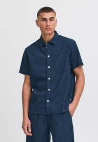 Camisa de manga corta azul marino con un tejido texturizado, cierre frontal con botones y un bolsillo en el pecho. Presenta botones blancos y un ajuste relajado.