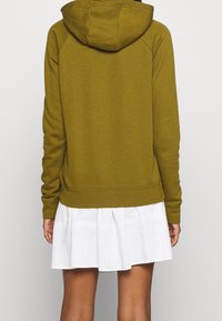 Person som bär en olivgrön hoodie och en vit plisserad kjol, sedd bakifrån mot en ljus, enfärgad bakgrund.