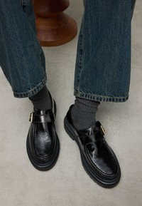 Zwarte gepolijste leren loafers met een afgeronde neus, gouden gespdetail en dikke zwarte rubberen zolen, gecombineerd met grijze sokken en spijkerbroeken.