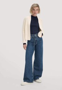 Blonde Frau, die eine cremfarbene Strickjacke über einem marineblauen Oberteil, hoch waistende weite blaue Jeans und weiße Sneaker trägt, steht mit einer Hand in der Tasche.