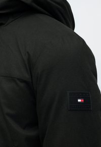 Čierna kapucňová bunda s textúrovanou tkaninou. Obsahuje nášivku s logom na rukáve s nápisom "TOMMY HILFIGER" a obdĺžnikovým dizajnom vlajky.