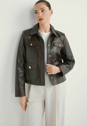 Kobieta w ciemnobrązowej skórzanej kurtce na białej bluzce i jasnobeżowych spodniach, stojąca na tle gładkiej jasnej ściany.