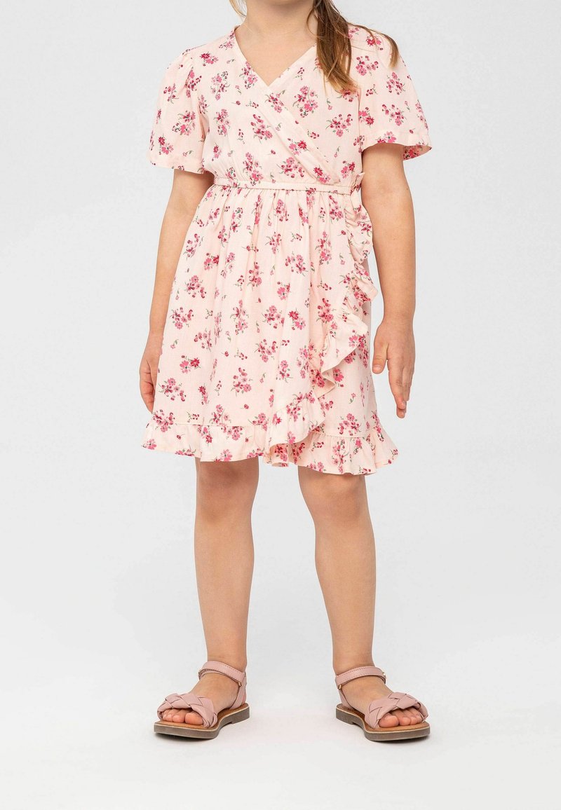 Robe fleurie rose avec manches courtes bouffantes, un design cache-cœur, un ourlet à volants et de petites fleurs dispersées sur toute la surface. Portée avec des sandales rose clair.