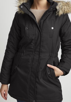 Parka noire isolée avec une capuche en fourrure, fermeture éclair à l'avant, boutons-pression, taille ajustable par cordon de serrage et poches à rabat, présentant une texture lisse.