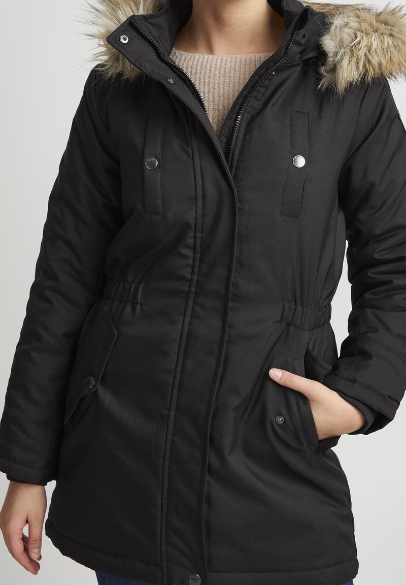 Parka noire isolée avec une capuche en fourrure, fermeture éclair à l'avant, boutons-pression, taille ajustable par cordon de serrage et poches à rabat, présentant une texture lisse.