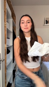 Kvinne med langt brunt hår iført en hvit kort topp og jeans holder et hvitt brettet plagg i et soverom med hyller og en seng.