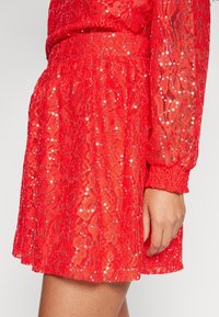 Jupe en dentelle rouge avec des motifs floraux et de petites paillettes dispersées, portée avec un haut en dentelle à manches longues assorti et un poignet élastique.