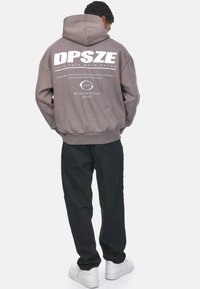 Grauer Kapuzenpullover mit großem weißen "DPSZE"-Logo auf dem Rücken, einschließlich Textdetails. Kombiniert mit schwarzen Hosen und weißen Sneakers.
