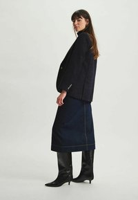 Blazer texturé bleu marine avec un col cranté, associé à une jupe en denim foncé et des bottes noires jusqu'aux genoux. La tenue présente des détails de couture visibles.