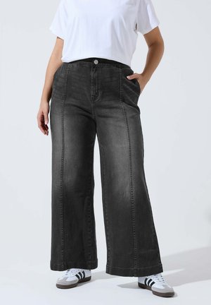 Person iført højtaljede sortvide jeans med brede ben, hvid t-shirt og hvide sneakers med sorte striber, stående med den ene hånd i lommen.