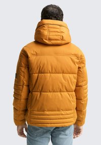 Orangene Pufferjacke mit Kapuze, gestepptem Muster und verstellbarem Kragen. Verfügt über eine glatte Textur und ein Patch auf dem linken Ärmel.