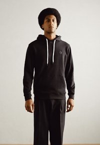 Sweat à capuche noir avec cordons blancs et poche kangourou, associé à un pantalon noir assorti. Le tissu semble doux, adapté à un style décontracté.