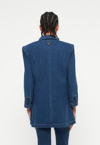Denim blazer v temno modri barvi z natančno zasnovo. Ima ovratnik z reverji, zapnene manšete in logotip na zadnji strani.