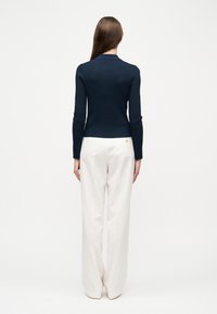 Γυναίκα με μακριά καστανά μαλλιά, ντυμένη με μπλε σκούρο μακρυμάνικο ριμπ τοπ και άνετο λευκό παντελόνι, που στέκεται γυρισμένη πλάτη σε λευκό φόντο.