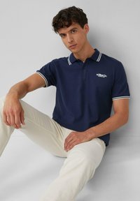 Marineblaues Poloshirt mit weißen Streifenakzenten am Kragen und den Ärmeln, aus einer Baumwollmischung, kombiniert mit hellbeigen Hosen, sitzende Pose.