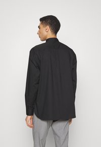 Camicia nera a maniche lunghe con colletto classico, tessuto di cotone liscio e design con yoke dritto sul retro, abbinata a pantaloni grigi.