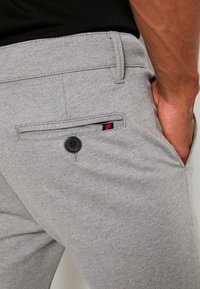 Gros plan d'une personne portant un pantalon gris clair avec une poche arrière noire boutonnée et la main reposant dans la poche avant.