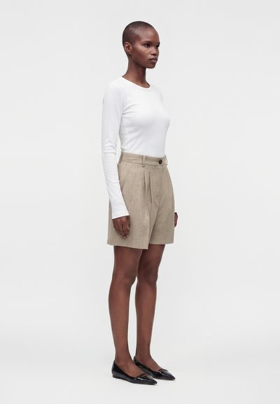 Femme debout portant un short plissé taille haute beige, un top à manches longues blanc et des ballerines noires pointues, sur un fond clair uni.
