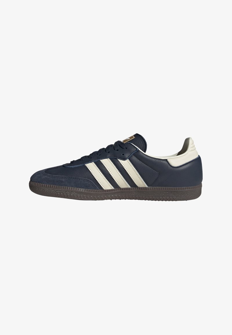Tumši zilas ādas sneakers ar trim bežām svītrām, teksturētu pirkstu daļu un brūnu gumijas zoli, ar kontrastējošu baltu papēža detaļu.