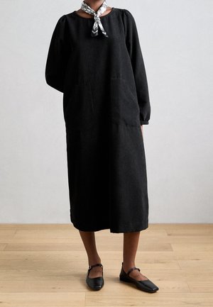 Žena v dlouhých černých texturovaných šatech s kapsami, s bílým vzorovaným šátkem na krku a černými botami Mary Jane na dřevěné podlaze.