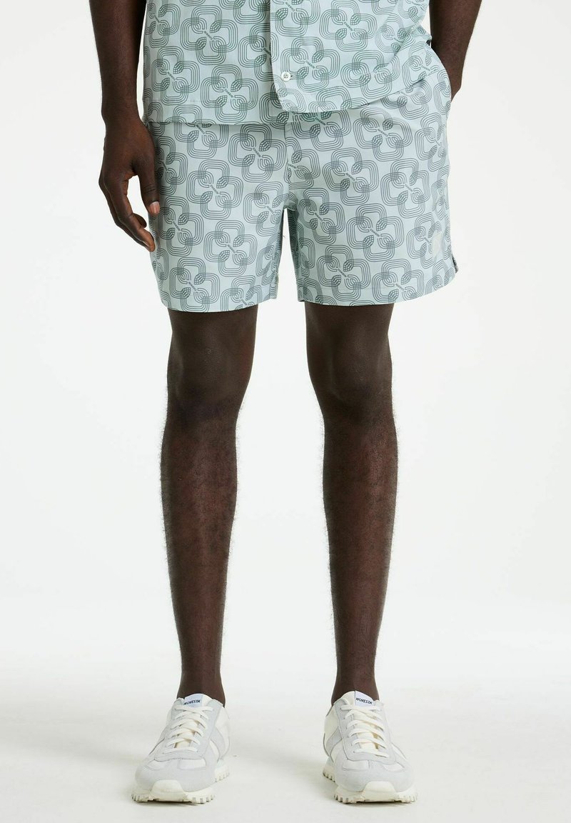 Homme portant un short vert clair et une chemise assortie à motif géométrique, debout en baskets blanches sur un fond blanc uni.