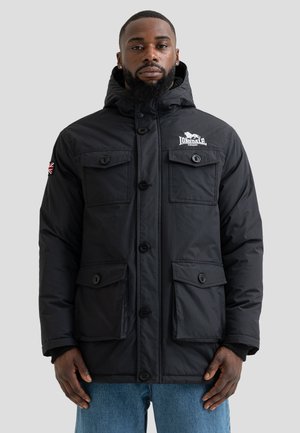 Homme portant une veste à capuche noire Lonsdale London avec poches avant et écusson du drapeau britannique sur la manche gauche, debout devant un fond uni.