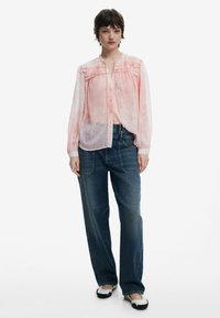 Blusa rosa chiaro, con bottoni e dettagli arricciati sulle spalle, abbinata a jeans scuri a gamba larga e ballerine bianco e nere.