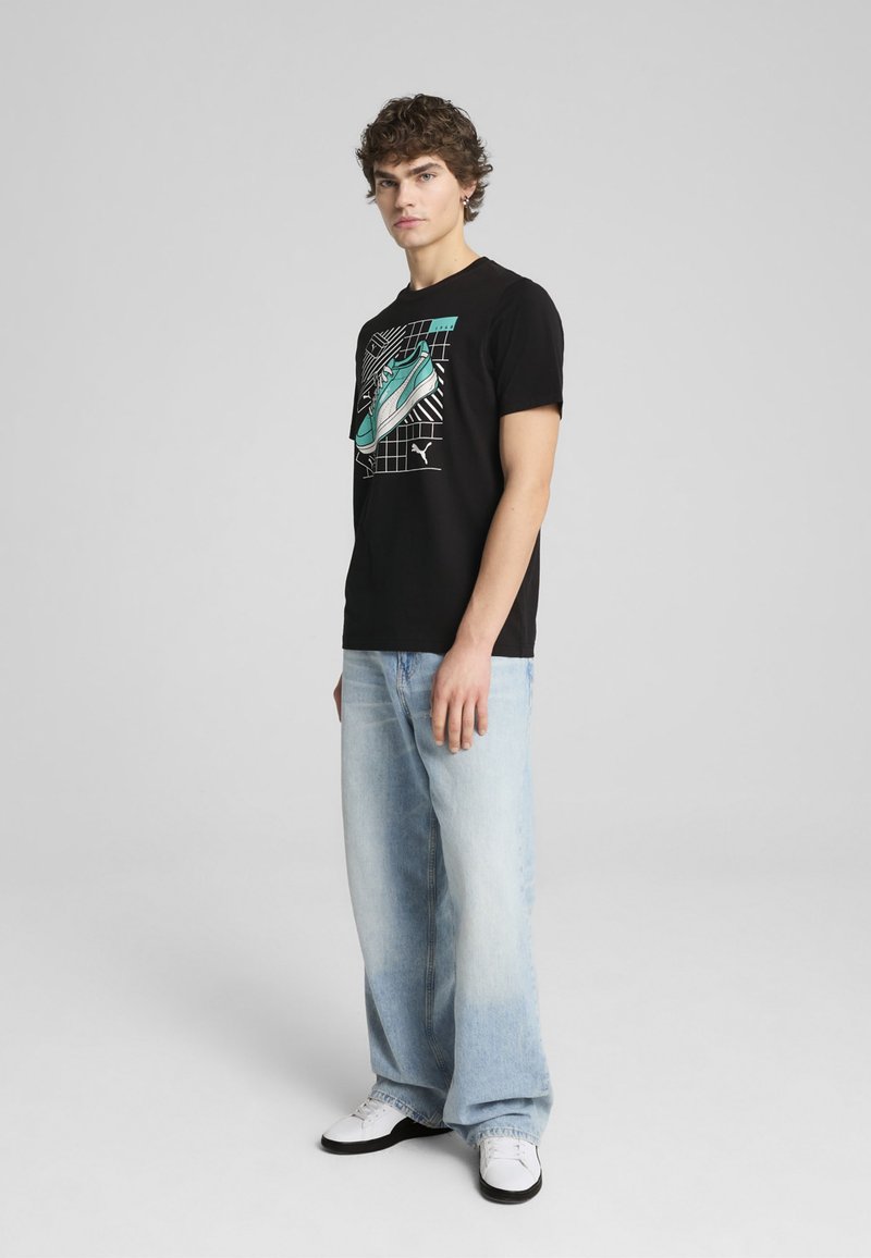 Sort t-shirt med et turkis sneaker grafik, parret med lyseblå brede jeans og hvide sneakers, mod en ensfarvet baggrund.