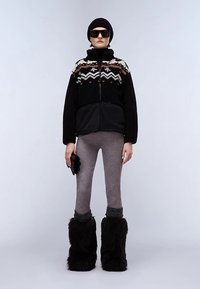 Czarna i szara kurtka zapinana na zamek z wzorzystą górną częścią, szare legginsy oraz czarne buty z sztucznego futra. Akcesoria obejmują czarną czapkę typu beanie i okulary przeciwsłoneczne.