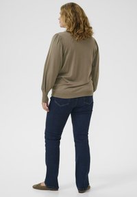 Vrouw met golvend haar staat met haar rug naar ons gekeerd, gekleed in een taupe langmouwen top, donkere spijkerbroek en bruine platte schoenen, tegen een witte achtergrond.