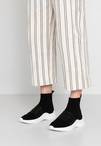 Černé slip-on tenisky s pleteným svrškem a texturovaným páskem. Mají bílou gumovou podrážku a ponožkový střih kolem kotníku pro bezpečné uchycení.