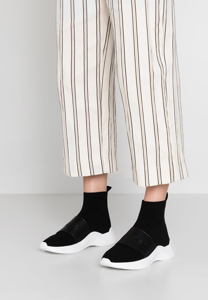 Černé slip-on tenisky s pleteným svrškem a texturovaným páskem. Mají bílou gumovou podrážku a ponožkový střih kolem kotníku pro bezpečné uchycení.