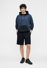 Fiatal férfi áll, sötétkék és fekete kapucnis felsőt, fekete rövidnadrágot és fekete sportcipőt visel, kezei a zsebében, fehér háttér előtt.