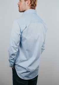 Chemise bleu clair à boutons, manches longues, texture douce, poignets pliés et ourlet incurvé, vue de dos.