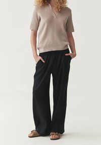 Haut beige en maille à manches courtes et encolure en V, associé à un pantalon noir à jambes larges. Le mannequin porte des sandales plates marron et a les mains dans les poches.